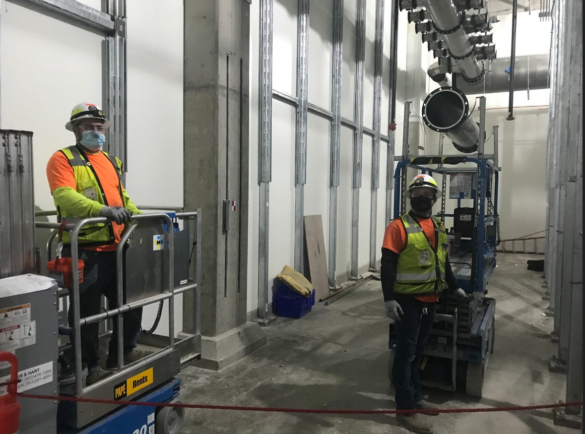 Two Harris & Hart workers in hard hats and masks on scissor lifts inside a facility, small-bore duct overhead
