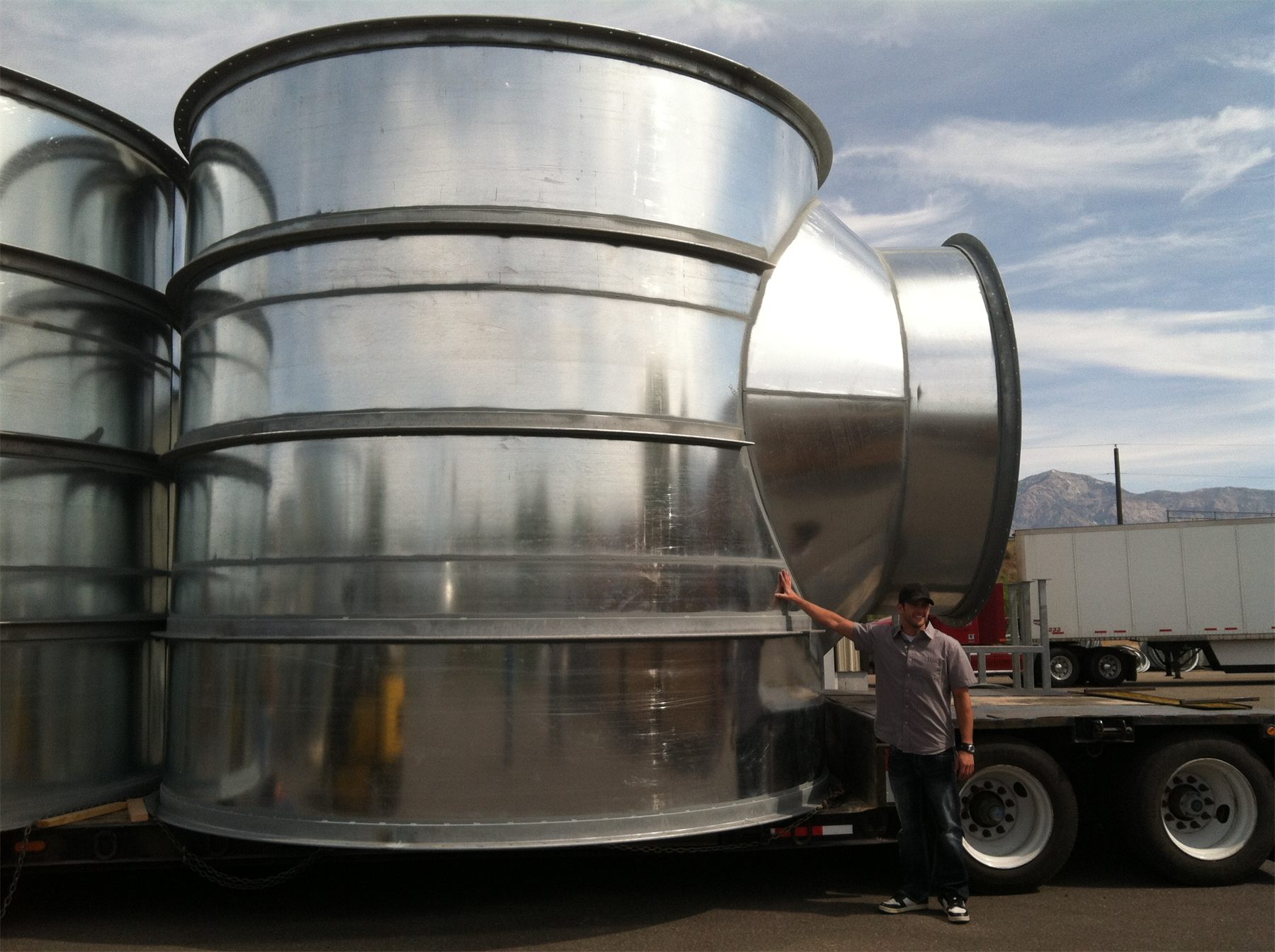 Worker standing beside a large galvanized round duct fitting on a trailer, for scale