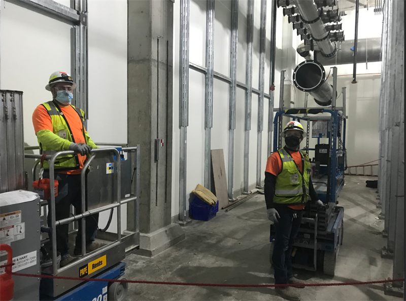 Two Harris & Hart workers in hard hats and masks on scissor lifts inside a facility