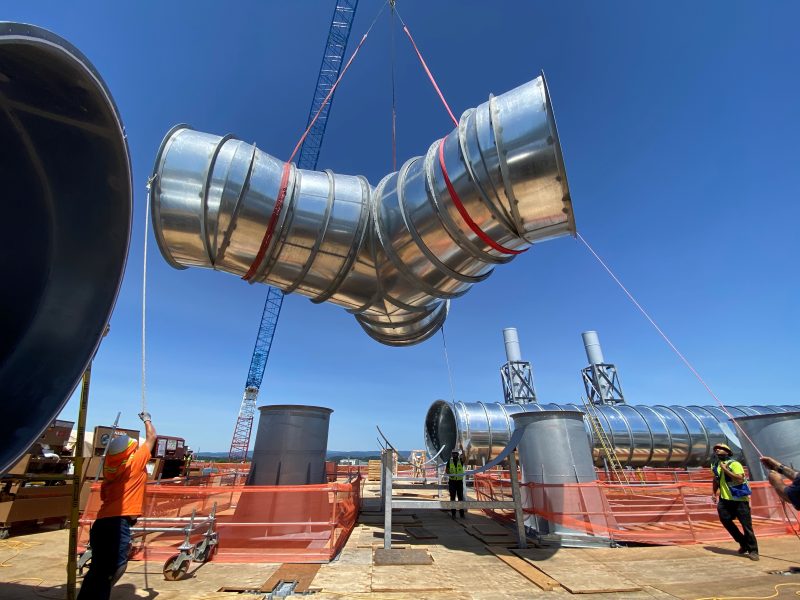Crew guiding a large round duct elbow on tag lines as a crane lifts it over a rooftop