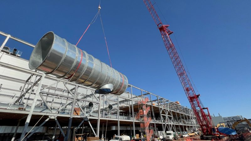 Crawler crane lifting a long section of large round duct onto a building's steel roof structure