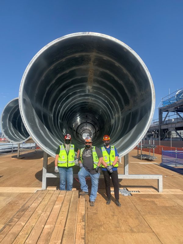 Three workers in hard hats and hi-vis vests standing in front of a very large round duct on a rooftop