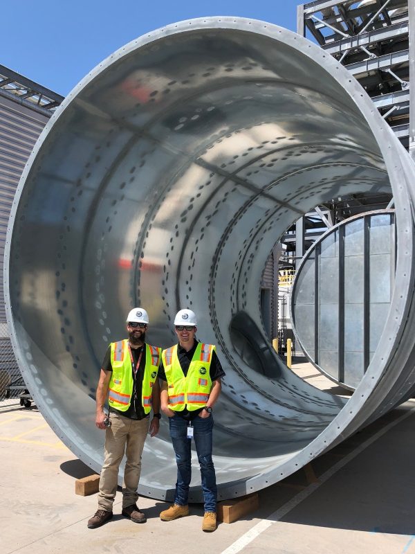 Two workers in hard hats and hi-vis vests standing in front of a very large round duct section